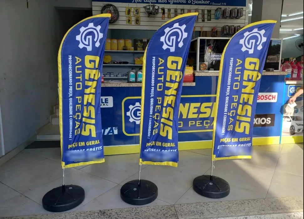 Três wind banners azuis e amarelos na entrada de uma loja de autopeças, produzidos pela Flamagraf